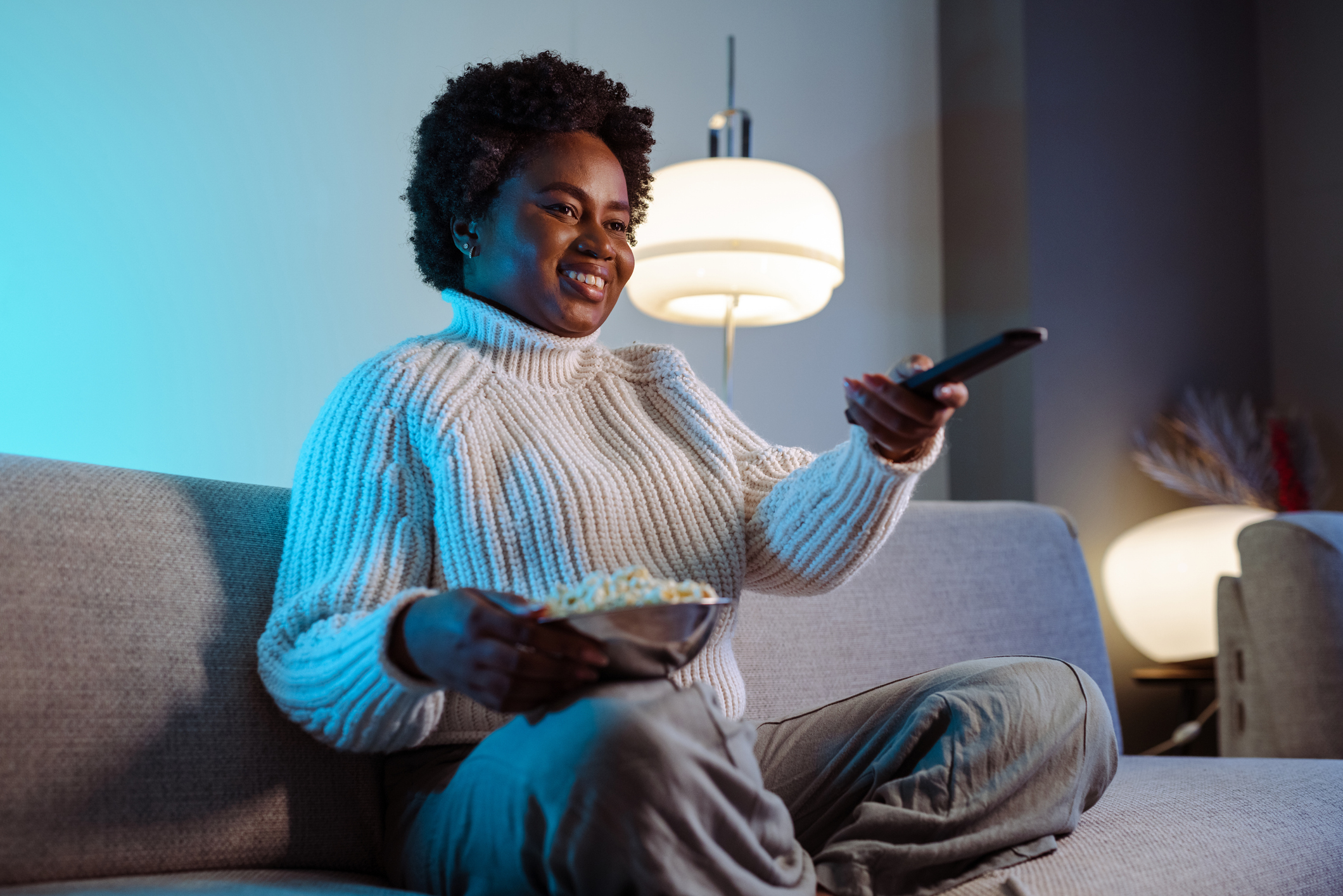 ALT Text: Cheerful smiling woman is watching TV and holding the remote control. Cozy night at home