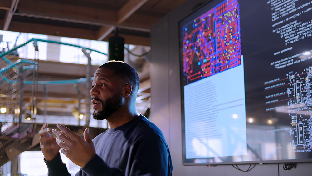 ALT Text: A man presenting his findings in a meeting with televisions behind projecting the data