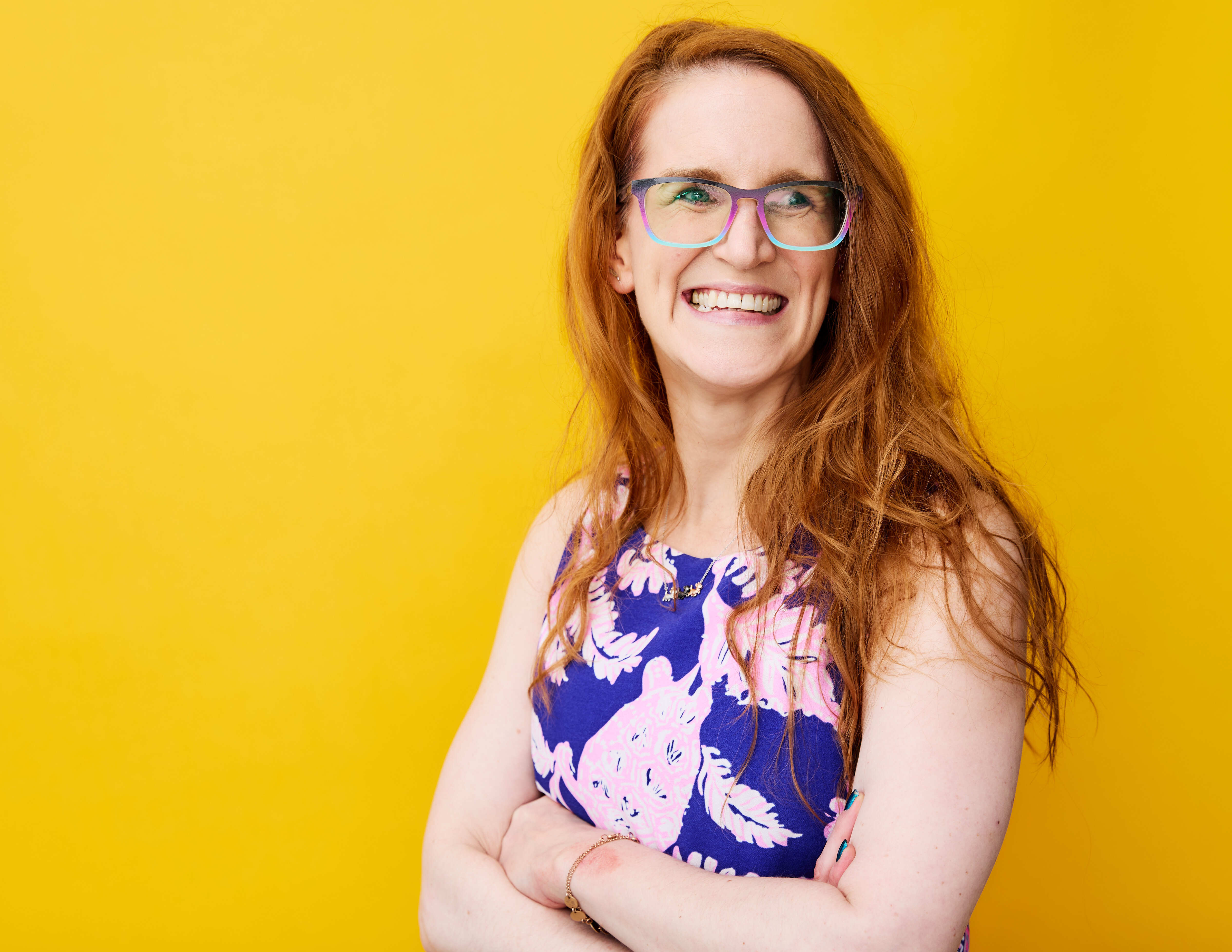 ALT text: a female ( Liz Schimidlin) with bright ginger hair and bright color glasses smiling. with a yellow background 